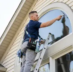 Professional window cleaner on a ladder using a squeegee to clean an exterior arched window, Spokane, WA.